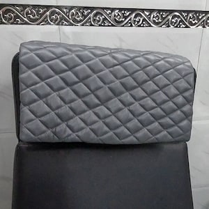 May include: A grey, quilted cover, possibly for a chair or appliance, is shown. The cover has a diamond pattern and is placed over a black surface. The background features a decorative silver trim.