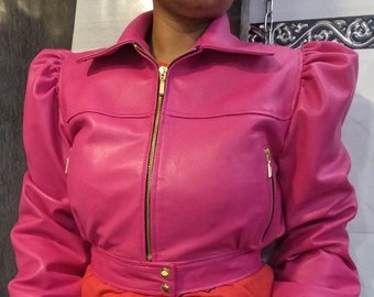 May include: A bright pink leather jacket with a zip-up front, puffed sleeves, and a collared neckline. The jacket has gold-colored zippers and buttons. The person is wearing a red shirt and blue jeans.