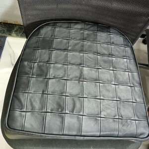 May include: Black leather chair cushion with a quilted design. The cushion is square shaped and made of a soft, padded material.
