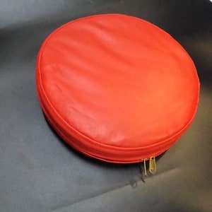 Puede incluir: Una bolsa redonda roja brillante con cremallera hecha de cuero. La bolsa está sobre una superficie gris oscuro.