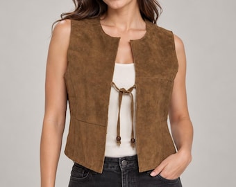 May include: A brown suede vest featuring a sleeveless design with a round neckline and a front tie closure. The vest has a textured finish and is worn over a plain white top paired with dark gray denim jeans, creating a layered appearance.
