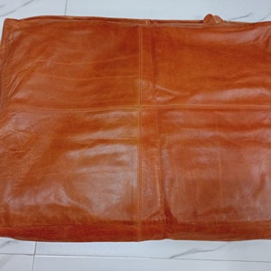 May include: A large, rectangular, brown leather tote bag. The bag has a stitched design and a handle. The leather has a distressed appearance with visible grain and variations in color, giving it a vintage look.