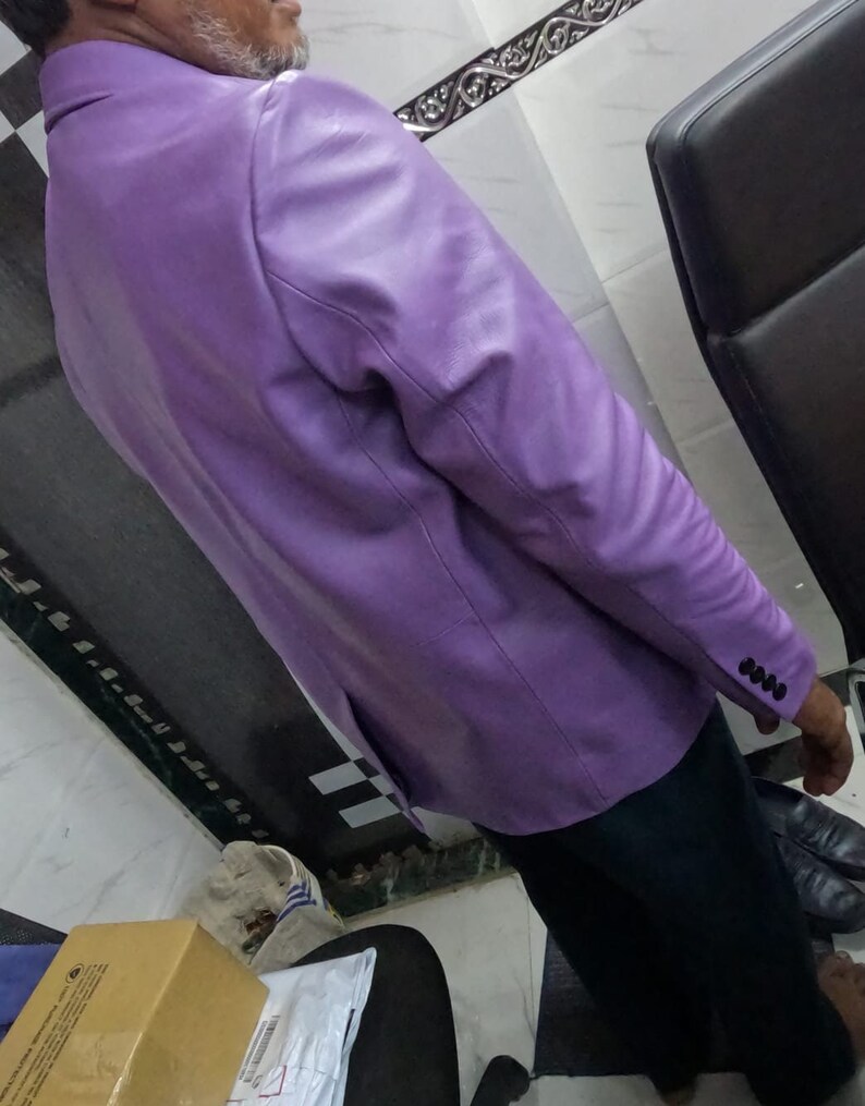 May include: A purple leather blazer with a buttoned cuff. The blazer is worn by a person standing in a room with a chair and a box.
