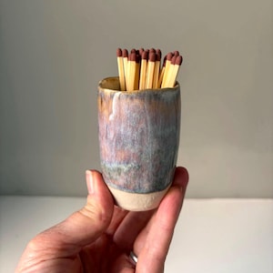 May include: A small, handmade ceramic match holder with a unique glaze in shades of blue, purple, and brown. The holder is filled with wooden matches, each with a red tip. The base of the holder is a natural, unglazed colour.