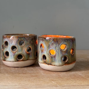 May include: Two cylindrical ceramic candle holders featuring a mottled glaze in shades of blue, green, and brown. Each vessel has a series of circular cutouts throughout the body, revealing a warm orange glow from within the interior space.