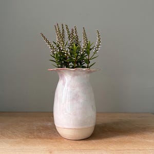 May include: A ceramic vase with a flared, ruffled rim and a smooth, glossy finish in shades of pale pink and white. The base is unglazed, showing a natural beige clay colour. A cluster of green stems with small white flowers is inside the vase.