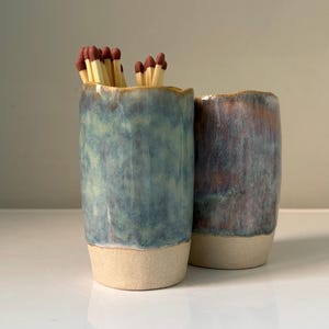 May include: Two ceramic match holders with a blue and green glaze. One holder contains matches with brown tips. The holders have a beige base and a textured surface. The holders are cylindrical and have a slightly irregular rim.