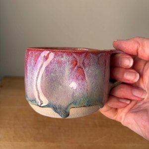 May include: A ceramic mug featuring a reactive glaze with blended hues of deep red, soft purple, and hints of blue that drip down towards the base. The bottom portion of the mug is made of unglazed, textured beige clay, providing a contrast in finish.