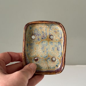 May include: A rectangular ceramic soap dish with four drainage holes. The dish has a speckled cream, blue, and purple glaze with a bronze-coloured rim. The dish is held by a hand.