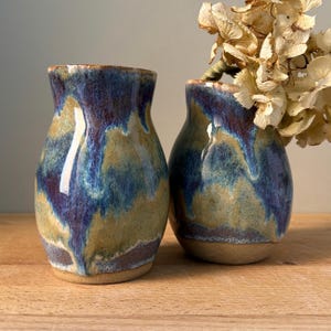 May include: Two ceramic vases with a blue, purple, and brown glaze. The vases are round and have a wide opening. They are sitting on a wooden surface.