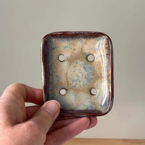 May include: A rectangular ceramic soap dish with rounded corners, featuring a mottled glaze in shades of tan, light blue, and dark brown. The dish has four circular drainage holes centred in the middle, with a raised rim around the edge.
