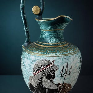 Può includere: Una brocca in ceramica con una finitura maculata color verde acqua e bianco e dettagli dorati. Il vaso presenta un motivo a greca attorno al collo e un'illustrazione in stile figure nere raffigurante una figura barbuta con un tridente.