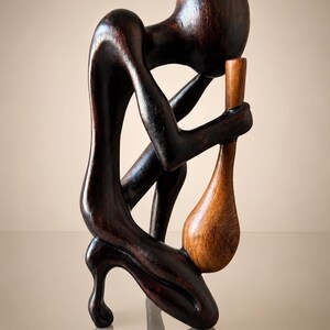 May include: A dark brown wooden sculpture of a kneeling figure with a smooth, rounded head and elongated limbs. The figure is leaning forward, holding a lighter, honey-colored wooden object shaped like a pestle or a teardrop in its hands.