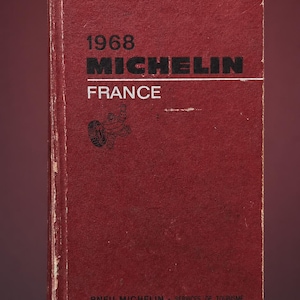 May include: A vintage 1968 Michelin France travel guide with a worn red cover. The front features black text reading 1968 MICHELIN, white text reading FRANCE, a small Michelin Man graphic, and address details at the bottom in small white print.