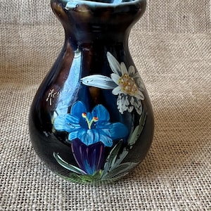 Könnte beinhalten: Eine dunkle, glänzende, bauchige Keramikvase mit handgemalten Blumenmotiven. Das Design zeigt einen markanten blauen Enzian und eine weiße Edelweißblüte mit grünen Blättern. Die Vase hat einen ausgestellten Rand und eine glatte, reflektierende Oberfläche.