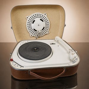 May include: A portable suitcase record player with a brown exterior and a white interior deck. It features a black circular turntable, a white tonearm, and a white circular speaker mounted in the lid. The text TEPPAZ CLED is printed near the tonearm.