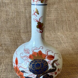 Può includere: Un vaso soliflore in porcellana bianca con collo stretto e base bulbosa. La superficie presenta motivi floreali dipinti a mano in tonalità blu scuro, arancione bruciato e oro, con delicate linee rosse che definiscono foglie e steli.