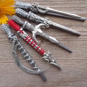 May include: An assortment of decorative letter openers in the shape of miniature swords and daggers, with intricate silver-colored metalwork and wrapped handles. One has a red handle. A yellow flower rests nearby on a wooden surface.