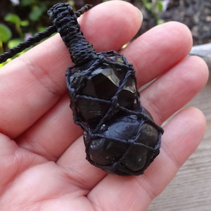 May include: A black macrame necklace featuring two dark, polished, irregular-shaped stones encased in a woven black cord netting. The pendant is suspended from a matching black cord, with a tightly knotted bail connecting the stone cage to the necklace.