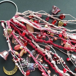 May include: A pink and red beaded and fabric tassel with bells, stars, and a crescent moon charm. The tassel is attached to a silver ring.