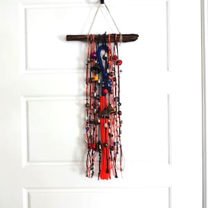 May include: A colorful macrame wall hanging with beads, bells, and tassels. The hanging is made with orange, red, blue, and white yarn and is attached to a driftwood branch.