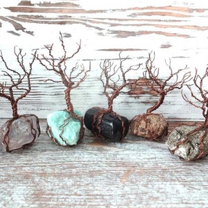 May include: Five handcrafted wire tree sculptures. Each tree features a copper-colored wire trunk and branches, rooted in a different colored stone. The stones include pink, turquoise, black, brown, and gray. The trees are displayed on a light wood surface.