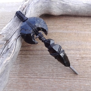 May include: A handcrafted obsidian axe with a dark, glossy finish. The axe head has a curved blade and is bound with black cord. The handle is also wrapped with cord, and the axe has a pointed tip. The axe is displayed on a wooden surface.