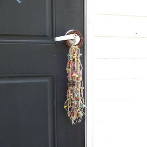 May include: A door with a beaded and bell door hanging. The door hanging is made of brown twine, colourful beads, and small bells. The door hanging is attached to a silver door knob.