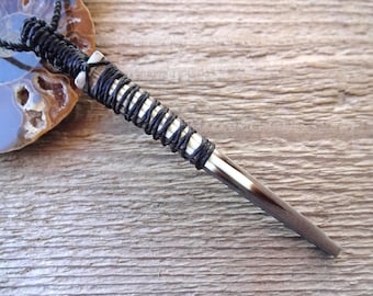 May include: A black cord wrapped around a long, thin, silver pendant with a black tip. The pendant is on a wooden surface.
