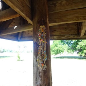 May include: A colourful beaded dream catcher hanging from a wooden beam in a gazebo. The dream catcher is made of brown string and has a variety of coloured beads, including red, orange, yellow, green, blue, and purple.