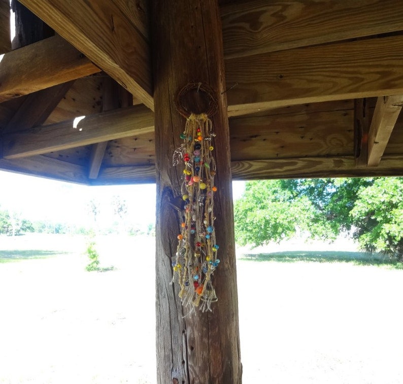 May include: A colourful beaded dream catcher hanging from a wooden beam in a gazebo. The dream catcher is made of brown string and has a variety of coloured beads, including red, orange, yellow, green, blue, and purple.