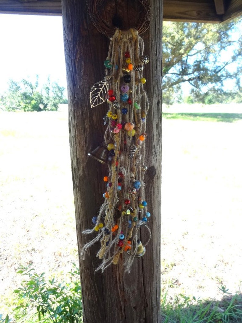 May include: A colourful beaded and bell wind chime with a leaf charm hanging from a wooden post. The wind chime is made of natural fibres and features a variety of beads in different colours and shapes.