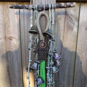 May include: A green, white, and brown macrame wall hanging with a hamsa hand charm and multiple bells. The hamsa hand charm is black with gold and purple accents. The bells are silver, gold, and purple.