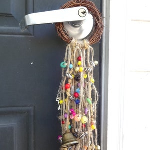 May include: A door with a wreath made of twigs and a door handle with a long strand of colourful beads and a small bell hanging from it.