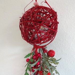 May include: A red woven sphere ornament with a cluster of red bells, berries, and green leaves. The sphere is made of intertwined twigs and has a small, silver-colored object inside. A decorative piece for the holidays.