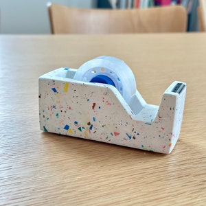 Könnte beinhalten: Ein weißer Klebebandabroller mit bunten Flecken. Der Abroller hält eine Rolle transparentes Klebeband. Der Abroller steht auf einem hellen Holztisch. Der Hintergrund umfasst einen Holzstuhl und ein Bücherregal.
