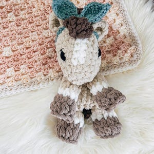 May include: A handmade crochet horse toy in beige, white, and brown, with a teal bow and brown flower detail. The toy is lying on a white fluffy surface next to a crocheted blanket in shades of pink, beige, and white.