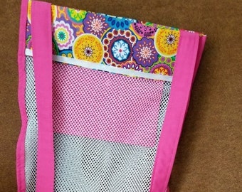 May include: A pink and white mesh beach bag with a colorful floral patterned top, featuring pink straps. The image also includes a yellow shovel and several seashells, suggesting a beach theme.