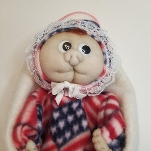 May include: A handmade doll wrapped in a white blanket. The doll's outfit is patterned with the American flag design in red, white, and blue. It has a round face, large eyes, and a bonnet with lace trim. A white bow is tied at the neck.