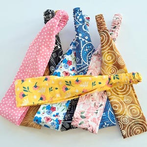 May include: A collection of colorful headbands with various patterns. The headbands include designs such as pink polka dots, black and white paisley, floral prints, and a yellow headband with a floral pattern. The headbands are made of fabric and have elastic bands.