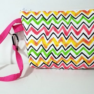 May include: A small, rectangular pouch with a white background and a colorful zigzag pattern in pink, green, yellow, and black. It has a pink strap with a black carabiner clip attached. The pouch has a zipper closure.