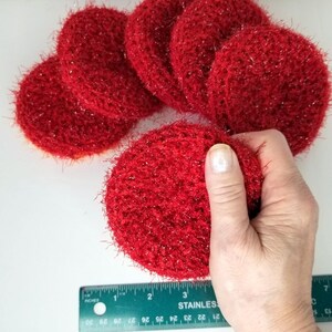 May include: Several round, red, crocheted scrubbies with a sparkly texture. One is being held by a hand. A ruler with inch measurements is in the foreground.