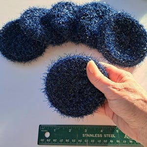 May include: Several navy blue, round, crocheted items are arranged in a curved line. One is held by a hand. A stainless steel ruler with inch measurements is in the foreground. The items appear to be approximately 4 inches in diameter.