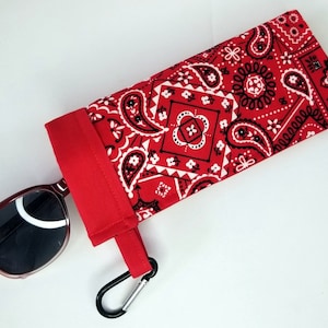 May include: A red bandana print sunglasses case with a black carabiner clip. The case is made of fabric and features a classic paisley pattern in white and black on a red background. Sunglasses are partially visible.