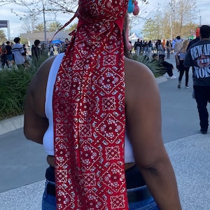 May include: A red durag featuring a white paisley design. The durag is worn on the head and extends down the back. The person is wearing a white top and blue jeans. A wristband is visible.