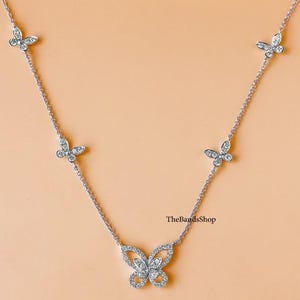 Puede incluir: Un collar de plata con cinco colgantes de mariposa de diamantes. El colgante de mariposa más grande está en el centro del collar, y los colgantes más pequeños están espaciados uniformemente a lo largo de la cadena. El collar está sobre un fondo naranja claro. El texto "TheBandsShop" es visible en la parte inferior de la imagen.