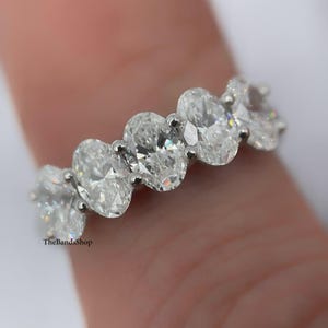 Puede incluir: Primer plano de un anillo de plata con múltiples diamantes de talla ovalada. Los diamantes están engastados muy juntos, creando un brillo continuo. El anillo está en un dedo, con el nombre de la marca "TheBandsShop" visible.