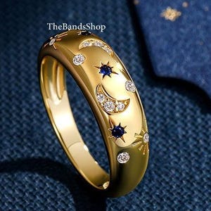 May include: A gold ring with a celestial design. The ring features crescent moons, stars, and small, round gemstones. The ring has a polished finish and is set against a dark blue background.