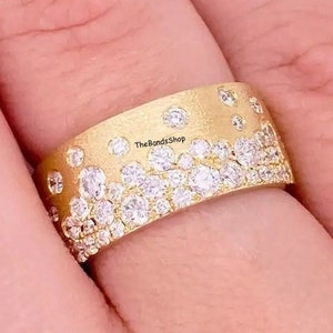 Puede incluir: Un anillo de banda de oro con un diseño de diamantes dispersos. El anillo está en un dedo y el texto "TheBandsShop" es visible en la banda.