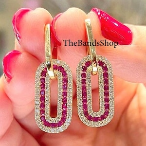 Puede incluir: Pendientes dorados con diseño ovalado. Los pendientes presentan una forma ovalada de doble capa, con una fila de pequeñas piedras preciosas rojas en el centro y un borde de piedras preciosas transparentes. Una persona sostiene los pendientes.